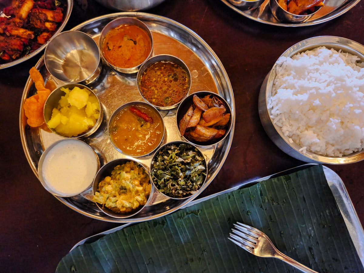 Thali vu du dessus : bols en inox garnis de currys, riz et condiments, posés sur une feuille de bananier, table en bois sombre.